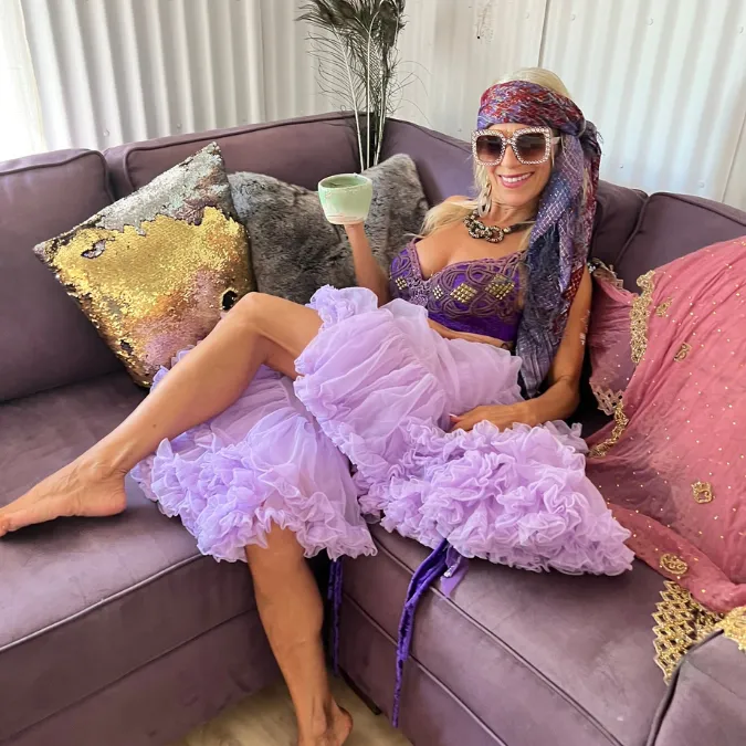 Staci relaxing barefoot on a lavender velvet sofa in a tulle skirt and headscarf, raising a cup of tea and smiling